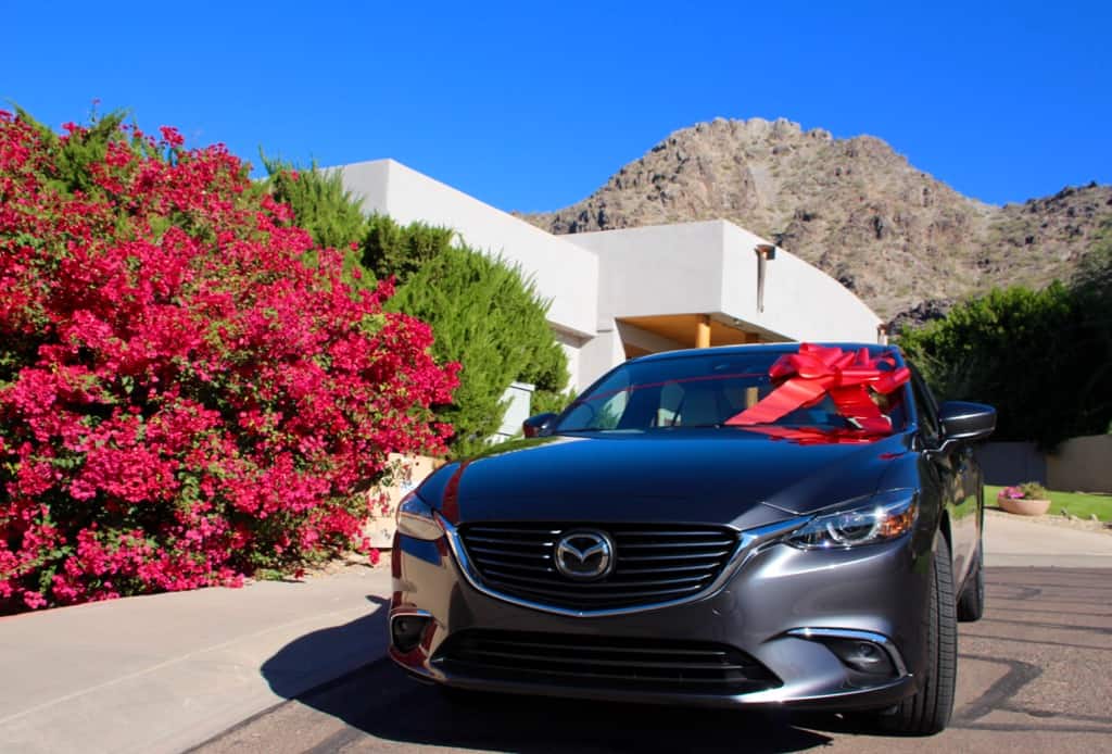 Holiday Road Trip Tips: Take a Trip, Travel Locally, silver Mazda6 with a white house and rock mountain, blue sky, giant red holiday bow